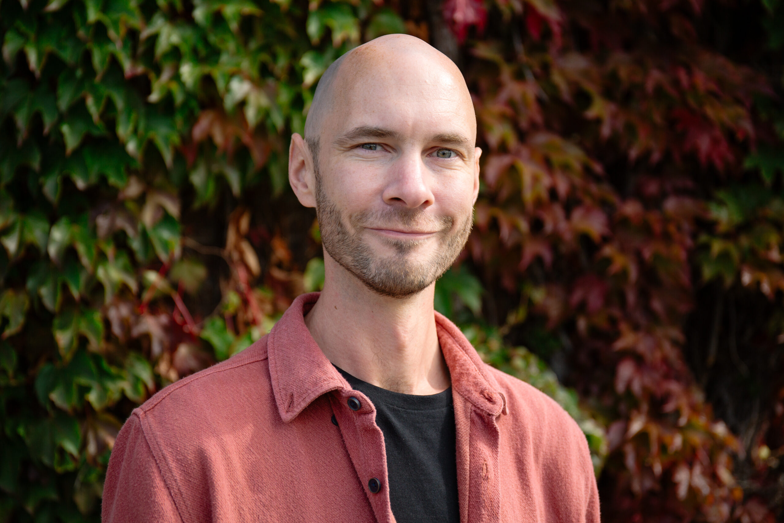 Photo of Dr Adam Alston, a member of the advisory board. He is stood outside in from of a hedge and wears a salmon-coloured flannel shirt.