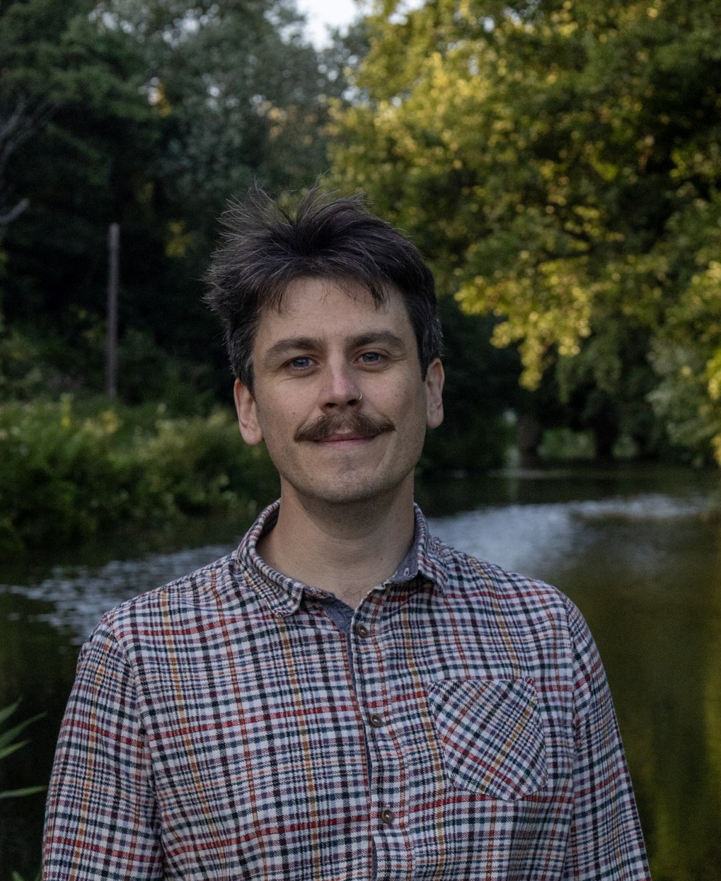 Photo of Cameron Dodds, a member of the advisory board. Cameron is stood outside in a woodland area wearing a checkered shirt in grey and black.