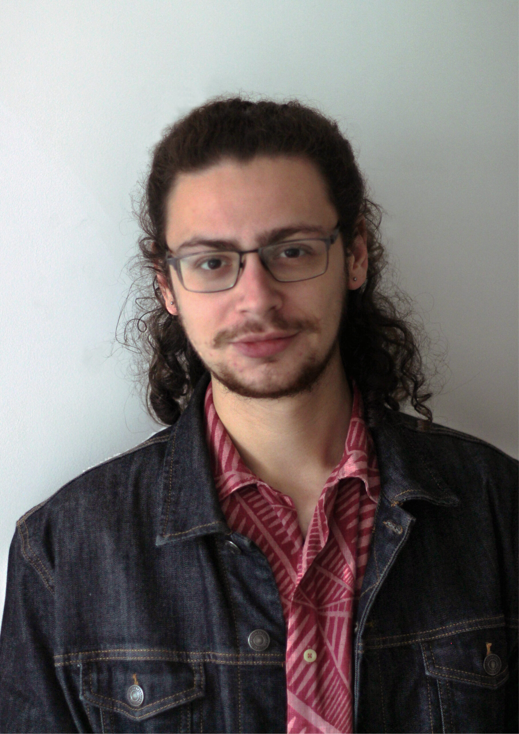 Photo of Noah Jay, the journal's editor. He has brown hair tied up in a bun and is wearing a denim jacket and red shirt.