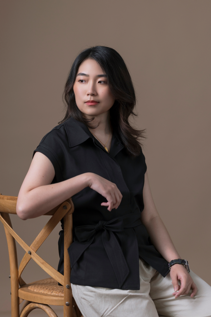 Photo of Zhui Ning Chang, a member of the editorial board. They are wearing a black shirt and are sitting sideways on a chair in front of a light brown background.