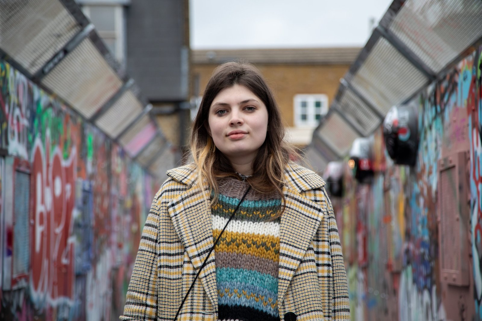 Headshot of Daisy Steinhardt, a member of the editorial board. Daisy is stood outside on a bridge. She is wearing a yellow patterned coat and a yellow and blue stripey jumper.